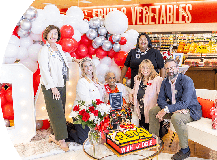 Various members of different organizations standing and sitting next to Romay Davis. They are all joyfully smiling. In front of the group there is a cake with the number '104'.