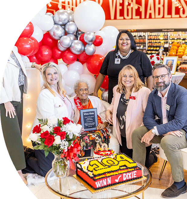 Various members of different organizations standing and sitting next to Romay Davis. They are all joyfully smiling. In front of the group there is a cake with the number '104'.
