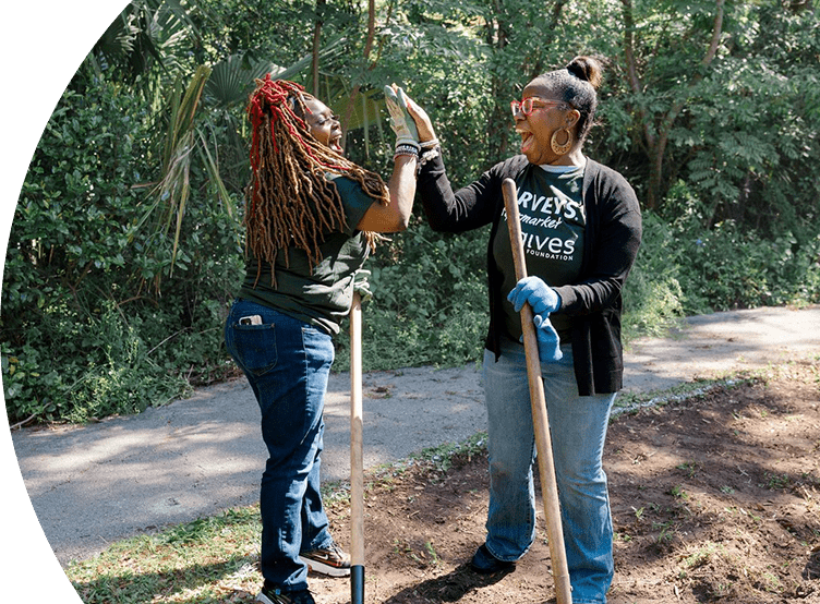Harveys Supermarket Give Foundation volunteers joyfully high-five one another. They are working with gardening tools.