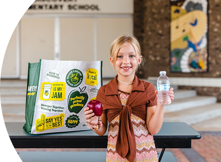 Girl standing while holding an apple and a water bottle. Behind her is a Harveys Supermarket reusable bag.