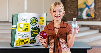 Girl standing while holding an apple and a water bottle. Behind her is a Harveys Supermarket reusable bag.