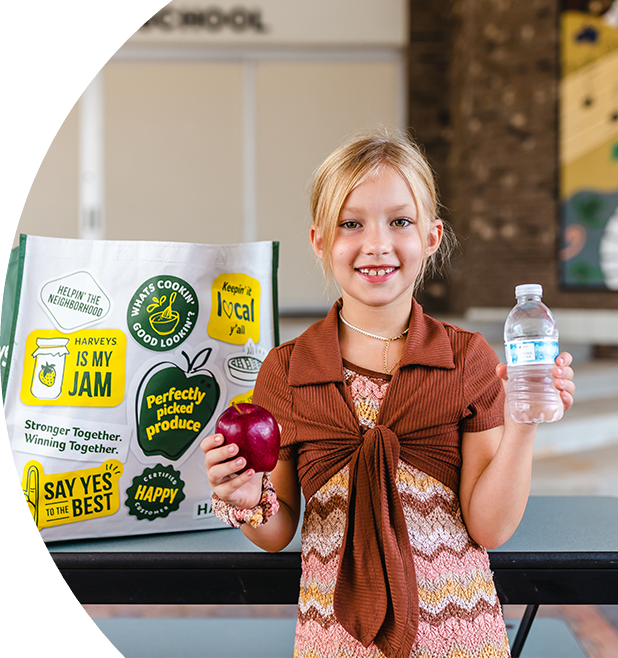 Girl standing while holding an apple and a water bottle. Behind her is a Harveys Supermarket reusable bag.