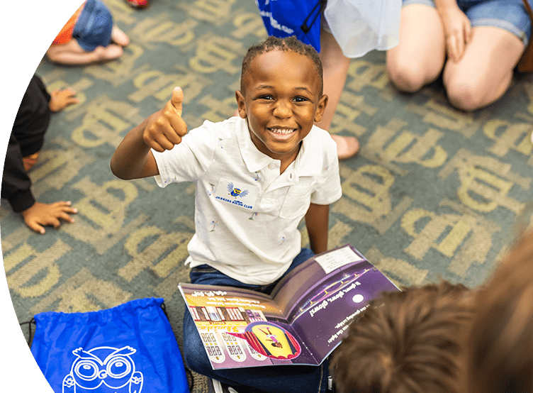 Smiling child giving a thumbs up. He is sitting criss crossed with a book on his lap.