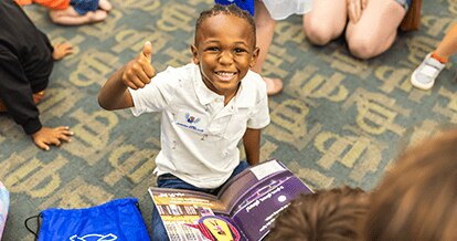 Smiling child giving a thumbs up. He is sitting criss crossed with a book on his lap.