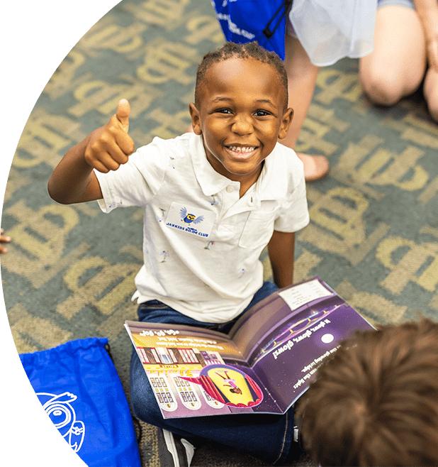 Smiling child giving a thumbs up. He is sitting criss crossed with a book on his lap.