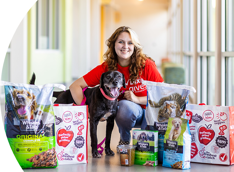 Pet food bags and Winn-Dixie re-usable bags set up in front of a woman kneeling and hugging a medium-sized dog. 