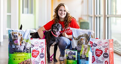Pet food bags and Winn-Dixie re-usable bags set up in front of a woman kneeling and hugging a medium-sized dog. 
