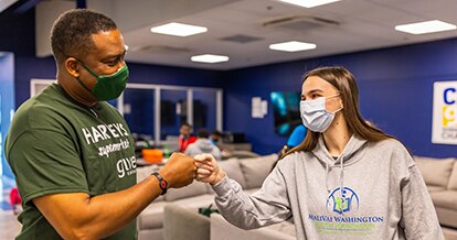 A young man in a Harveys Supermarket SEG Gives Foundation shirt and young woman in a MaliVai Washington grey sweatshirt wearing masks fist bumping.